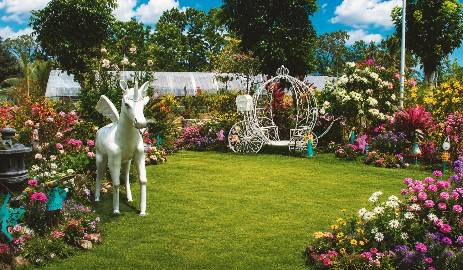 A white fairy-tale carriage display surrounded by blooming pink flowers and a unicorn statue in the whimsical garden area of Wondellea